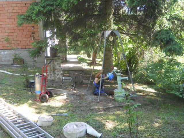 Tronožac sa čekrkom iznad otvorenog kopanog bunara u dvorištu, sa pumpom, crevima i merdevinama pored.