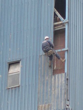 Radnik sa užeta, u pojasu i sa šlemom, radi na limenoj fasadi industrijskog objekta.